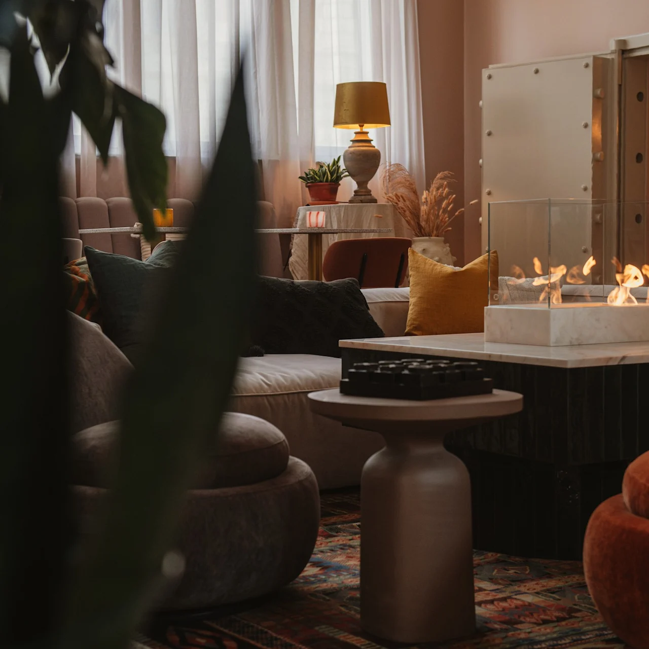 A glass-encased tabletop fire burns on a marble surface in a cozy living room, surrounded by plush sofas, warm pillows, an elegant lamp, and a large plant.
