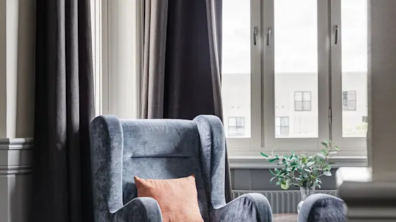 A blue armchair with an orange pillow stands near tall windows, framed by dark curtains in a carpeted room. A small plant sits on a table beside it.