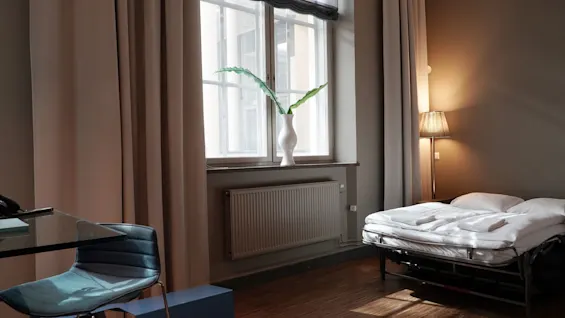 A white vase with green leaves stands on a windowsill. Nearby, a bed and lamp sit in a room with beige curtains and wooden flooring.