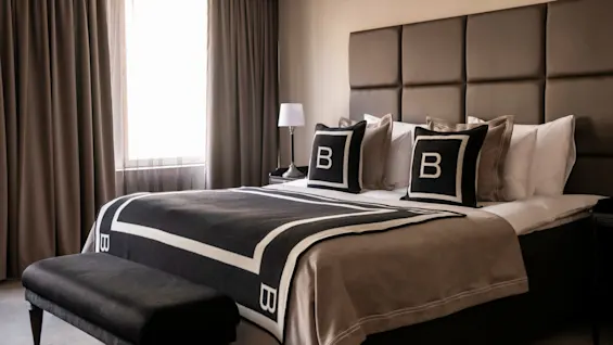 En omhyggeligt redt seng med puder mærket med "B" og et mønstret tæppe står i et blødt oplyst rum, flankeret af en lille bordlampe, med gardiner der indrammer et vindue.