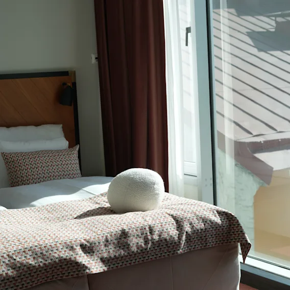 Hyggelig hotelseng med mønstret dyne og rund pude ved et solrigt vindu