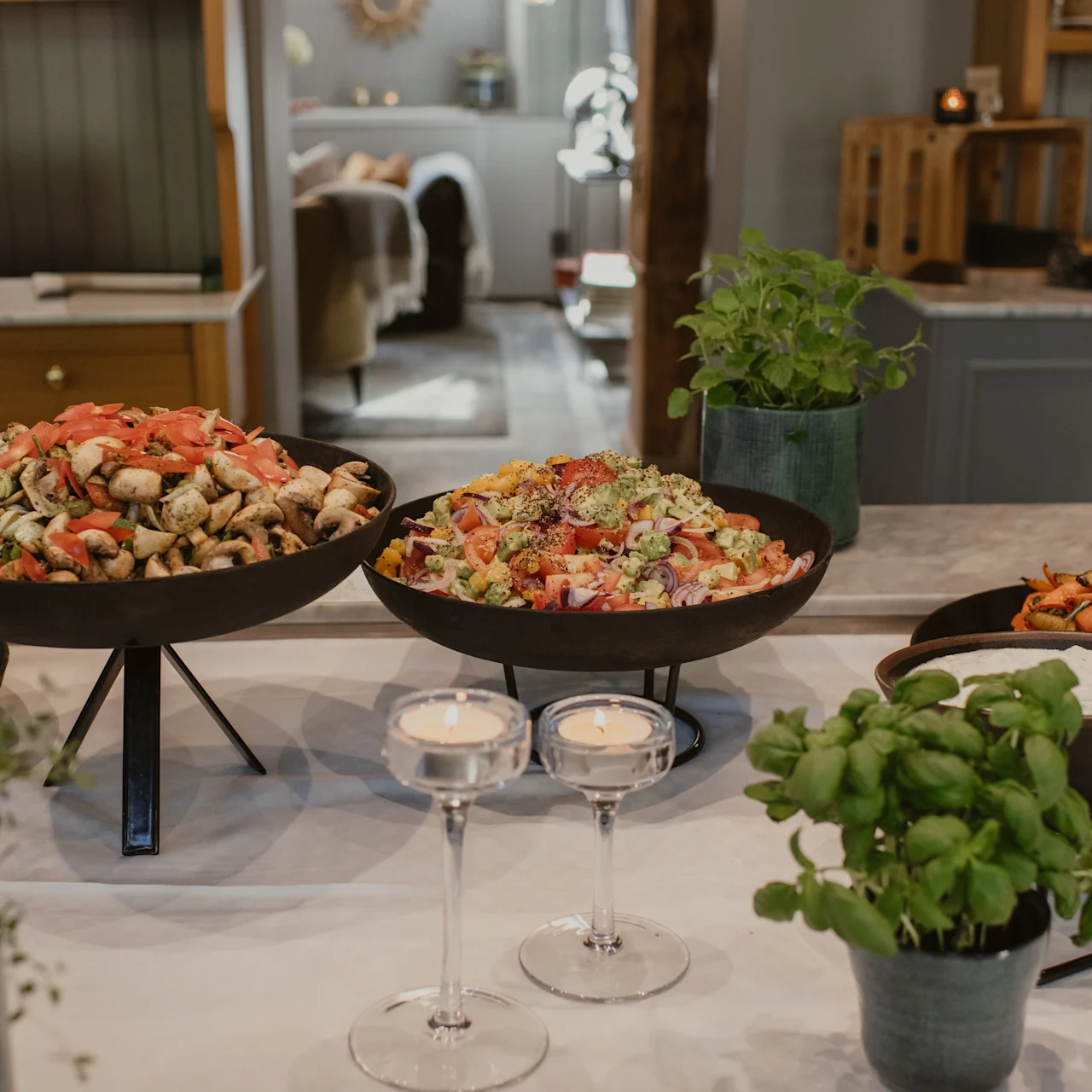 En buffé med olika sallader, rostade grönsaker och örter på en bänkskiva i ett mysigt, varmt belyst rum med suddiga bord och hyllor i bakgrunden.