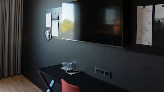 En väggmonterad TV ovanför ett mörkt skrivbord med två dricksglas i ett rum med svarta väggar. I närheten är papper uppsatta på en anslagstavla bredvid TV:n.