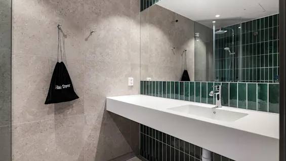 Moderne bad med hvit vask, stort speil og dusj i speilbildet. En svart pose merket 'Hair Dryer' henger på veggen.