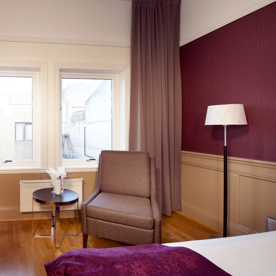Et hotellrom interiør med en stor seng, et skrivebord med en fjernsyn, en stol, og vinduer som ser ut mot en bygning.
