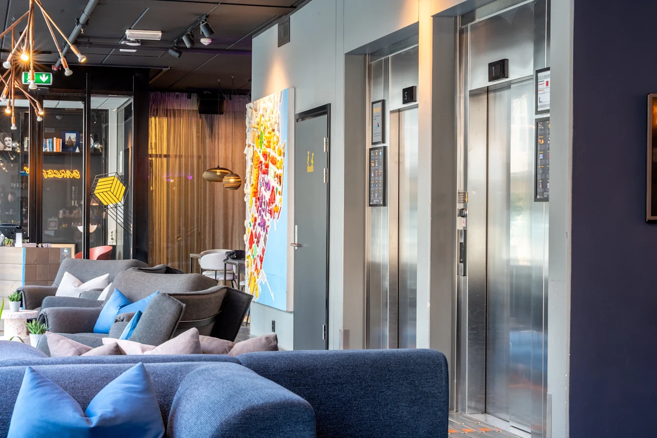 To lukkede elevatorer i en moderne lounge, omgivet af farverige sofaer og abstrakt kunstværk, med hylder og belysning i baggrunden.