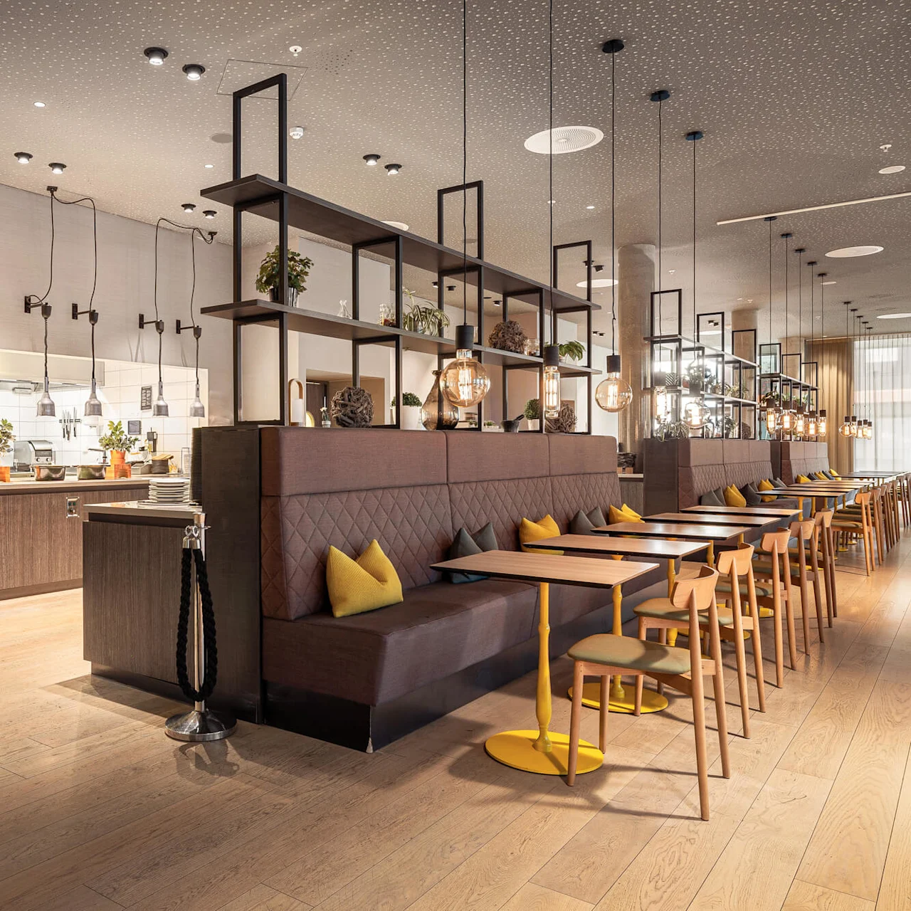 Restaurant interior features brown seating, yellow cushions, and wooden tables aligned in rows. Overhead, modern pendant lights illuminate. Neon sign reads "FOOD PICK-UP POINT". Cozy and inviting ambiance.