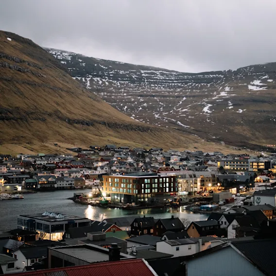 En naturskön vy över en kuststad i skymningen, med upplysta byggnader som speglar sig i vattnet och ett stort berg i bakgrunden.