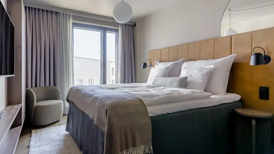 A neatly made bed with white linens and a gray throw blanket sits in a modern room, featuring a padded headboard, bedside lamps, a rug, and a window with curtains.