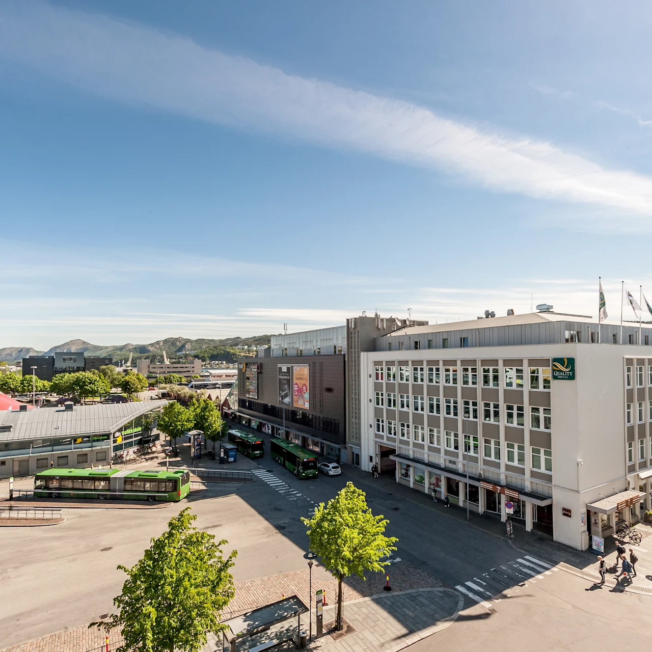 Suuri hotellirakennus, jossa on "Quality" kyltti, sijaitsee bussiterminaalin vieressä, jossa on vihreitä busseja. Tilanne on aurinkoinen ja kaupunkimainen, taustalla vuoria.