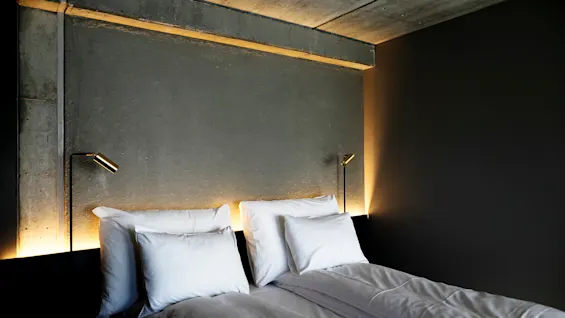Seng med hvide puder og lagner, ledsaget af to læselamper, placeret mod en minimalistisk betonvæg, der skaber en moderne og hyggelig atmosfære.