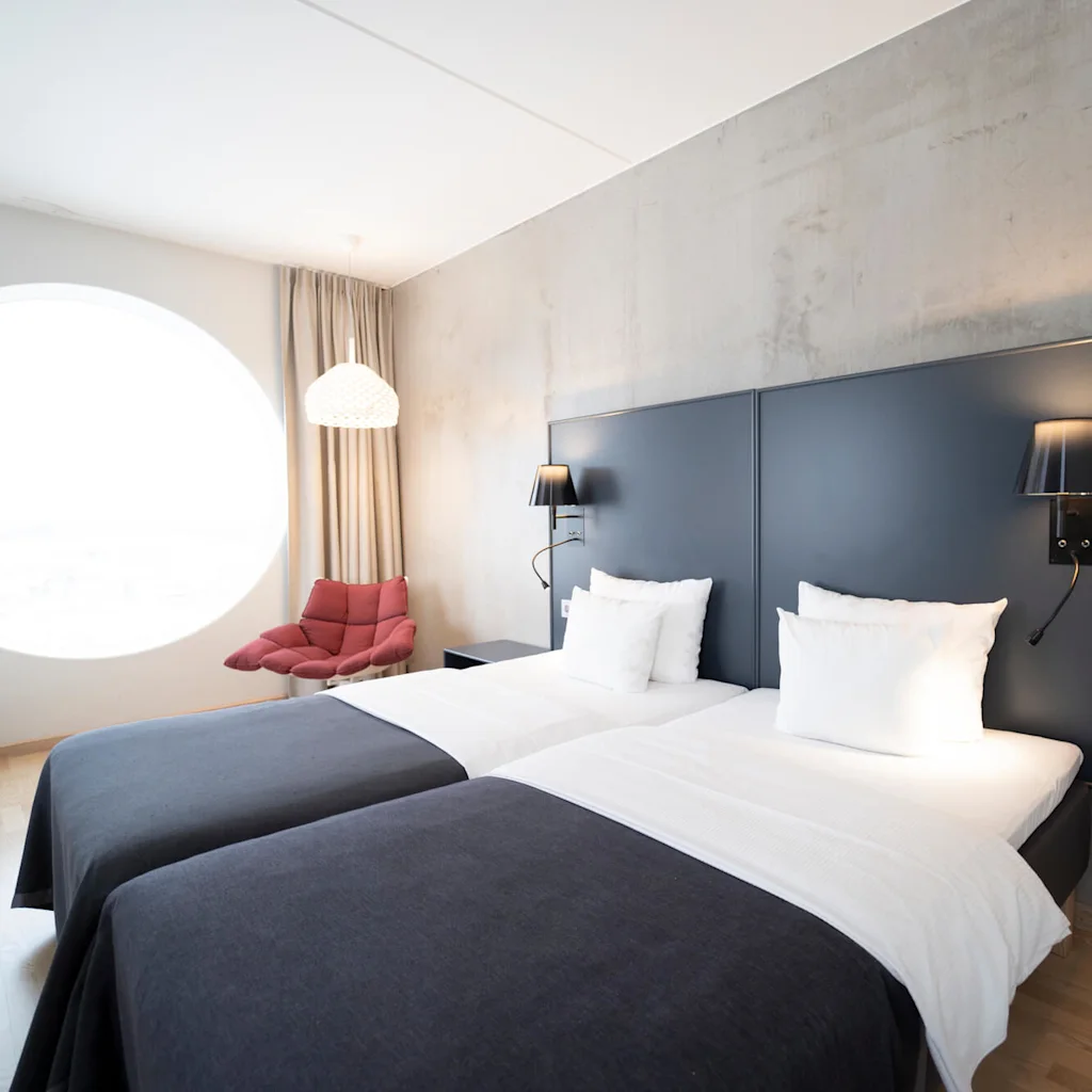 Twin beds with white linens stand against a gray wall, accompanied by black wall lamps. A red chair sits near a large circular window, warmly lit by a hanging lamp.