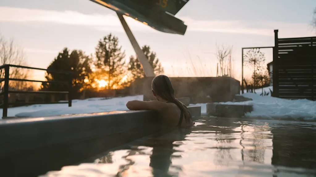 En person slapper af i et udendørs hot tub under solnedgang, omgivet af sne. En stor flyvemaskineskulptur med "35" står i nærheden.