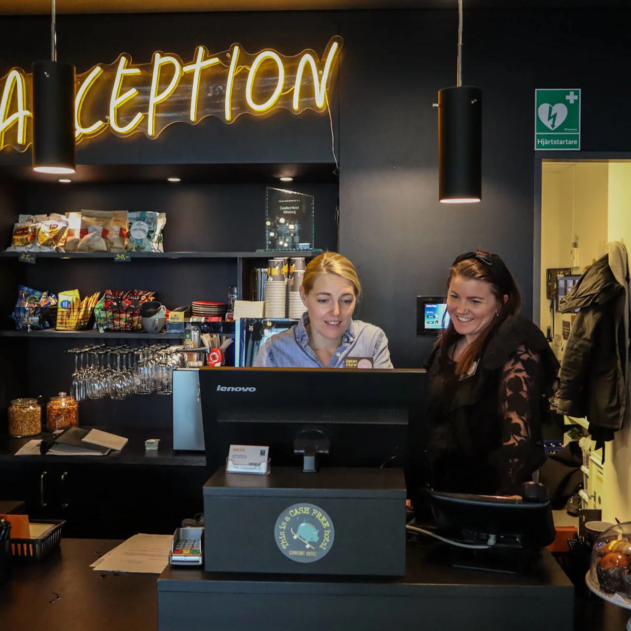 Två personer står bakom en receptionsdisk, fokuserade på en dator. Hyllor med snacks och glas i bakgrunden. En neonskylt lyder "BARCEPTION". En skylt läser "Hjärtstartare."