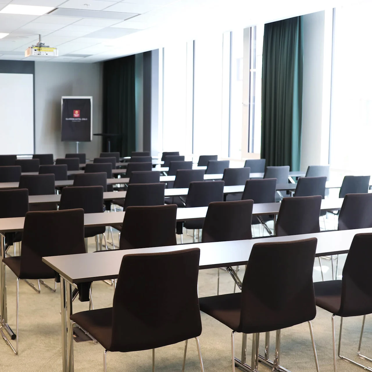 Empty conference room features rows of black chairs and tables facing a large white projection screen. The room is well-lit with natural light from tall windows.