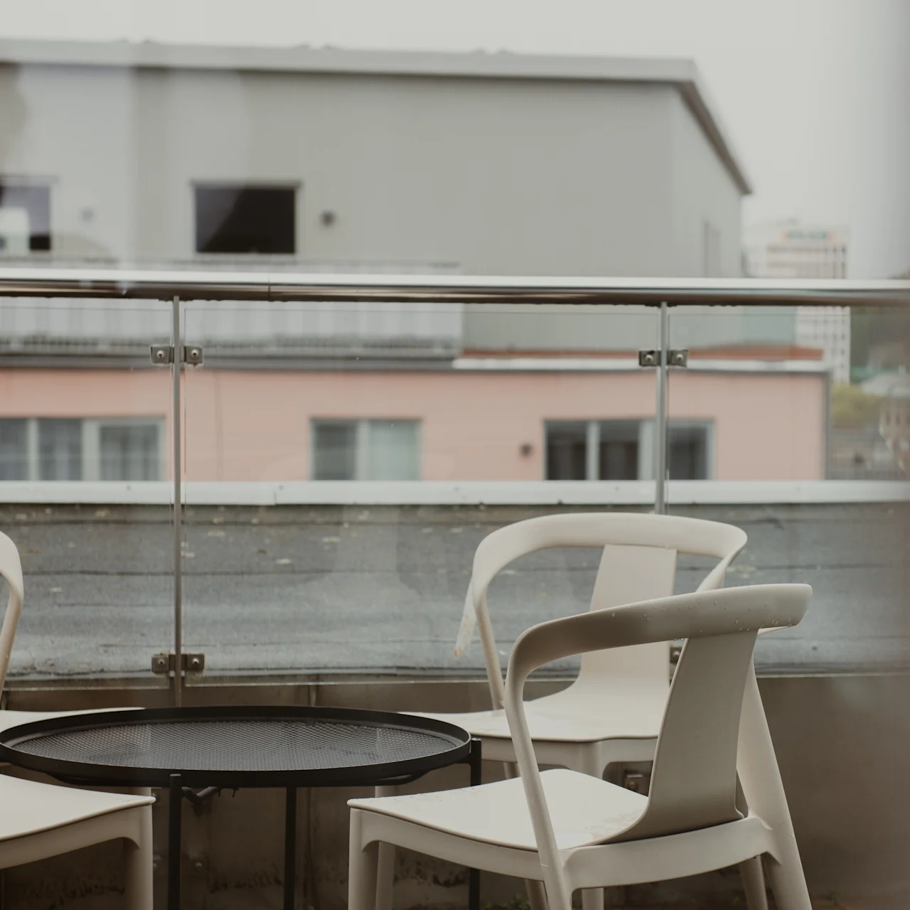 Four white chairs surround a round black table, situated on a balcony with glass railing, overlooking distant, blurred buildings under a cloudy sky.