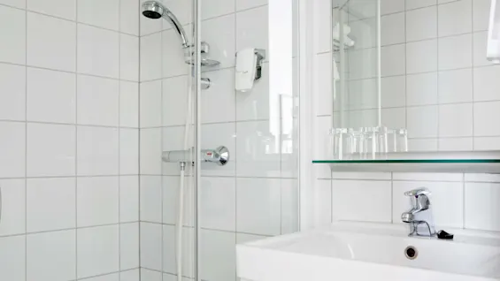Suihkupää kiinnitetty kaakeloituun seinään vierekkäin pesualtaan kanssa, jossa on kromihana. Laseja on sijoitettu hyllylle lavuaarin yläpuolelle siistissä, modernissa kylpyhuonetilassa. Teksti: "lux".