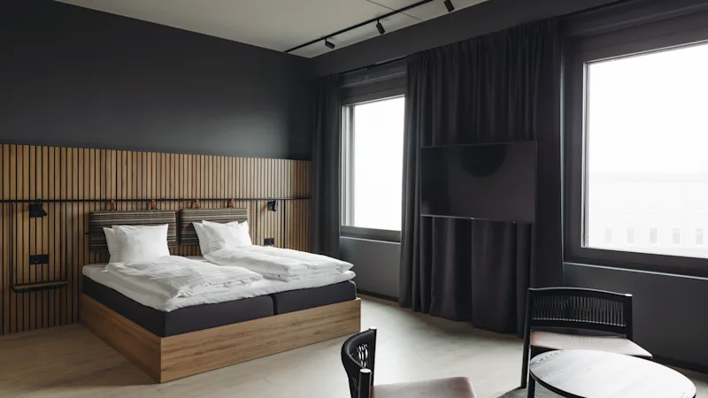 Et moderne hotelværelse med en stor seng, trægavl, mørke vægge og et siddeområde ved vinduet.