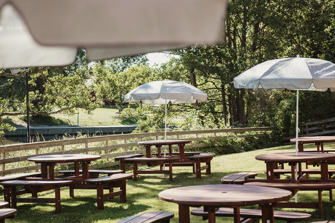 Utomhus picknickområde med träbord och parasoller en solig dag, intill ett lugnt vattendrag och lummiga gröna träd.