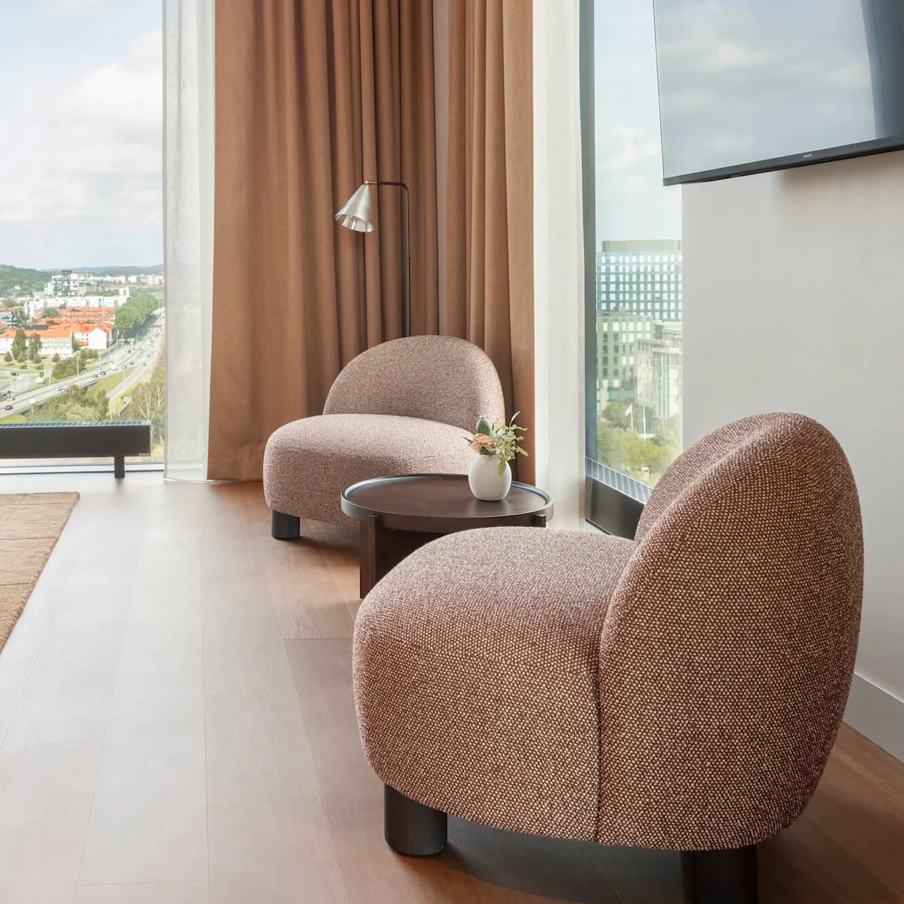 Två bruna loungestolar står bredvid ett litet träbord i ett modernt rum med stora fönster och beige gardiner. En väggmonterad TV är synlig.