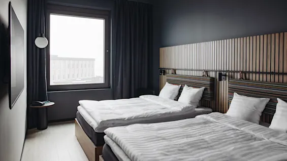 Two neatly made beds, topped with white linens, rest in a modern room. A large window reveals an urban building view, surrounded by dark walls and curtains.