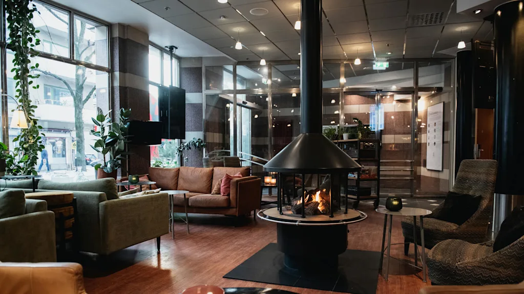 En hyggelig stue med en central pejs omgivet af sofaer og stole. Store vinduer giver naturligt lys, og stearinlystændte borde tilføjer varme. Potteplanter pynter det moderne rum.