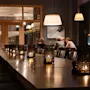 Stearinlys lyser opp et langt trebord, og skaper en varm atmosfære i en moderne restaurant. Folk samtaler i bakgrunnen under store pendellamper.