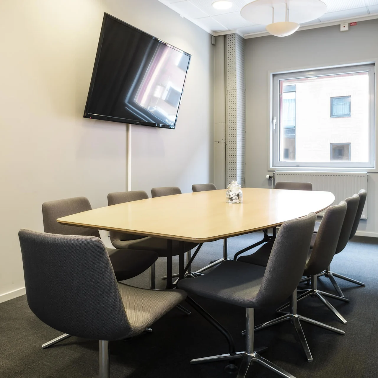 A large wooden conference table is surrounded by eight gray chairs, situated in a meeting room with a wall-mounted TV and a window with curtains.