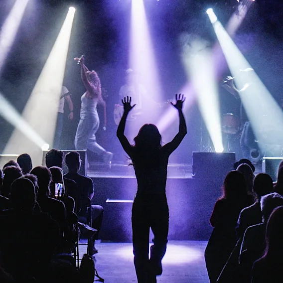 En konsertscene med artister belyst av skarpe lyskastere, og en person i silhuett som strekker hendene i været foran publikum.