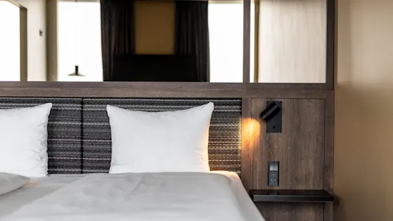 Seng med hvite puter og laken; leselampe over nattbord lyser varmt i et moderne, trepanelt hotellrom. Et vindu lyser mykt opp rommet bakfra.