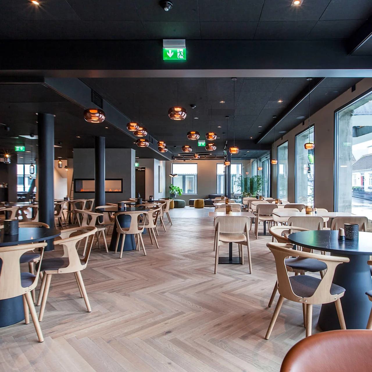 Stoler omgir runde bord i en moderne, romslig restaurant med tregulv. Store vinduer gir rikelig naturlig lys. Moderne pendellamper henger fra taket. Et utgangsskilt sier "Nødutgang."