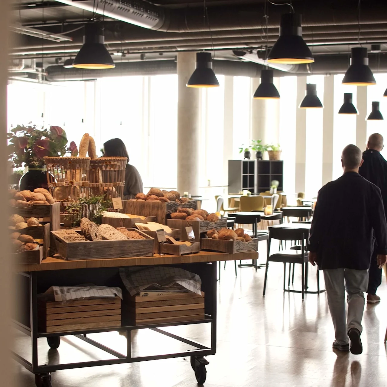 Bröddisplay på tralle av tre, med ulike brød og kurver; folk går forbi, plassert i en godt opplyst kafé med hengende lamper og tregulv.