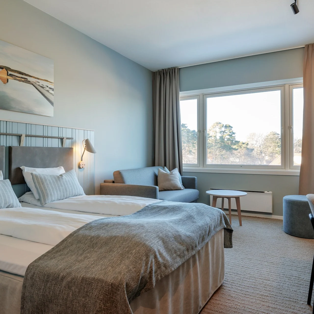 A neatly arranged hotel room features a double bed with gray bedding, a small desk, and a seating area by a large window. A landscape painting decorates the wall.
