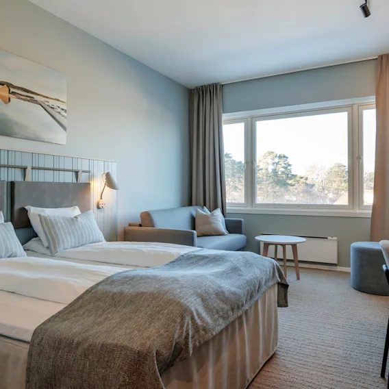 Et pent innredet hotellrom har en dobbeltseng med grått sengetøy, et lite skrivebord og en sittegruppe ved et stort vindu. Et landskapsmaleri pynter veggen.