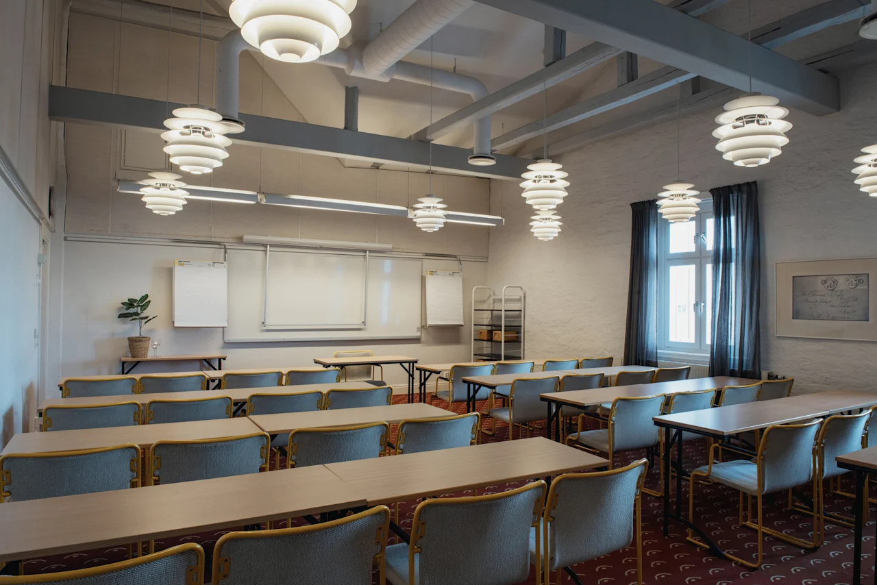 Ett rymligt konferensrum med flera bord och stolar, whiteboards och dekorativa taklampor, förberett för en sammankomst.