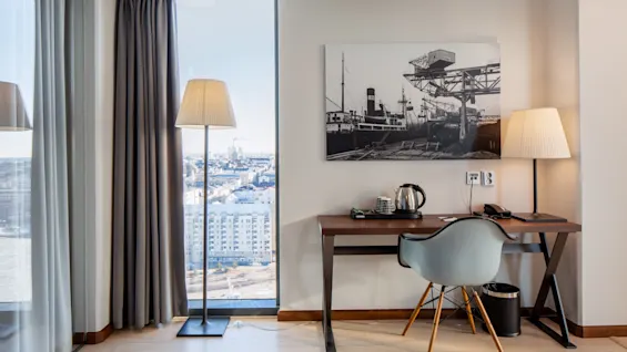 Et skrivebord med en blå stol vender mod en væg i et rum. På skrivebordet er der en kedel og telefon. En gulvlampe står ved et vindue med byudsigt og gardiner.