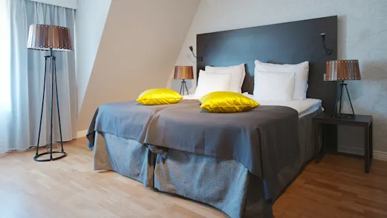 A neatly made bed with grey and yellow bedding in a bedroom, featuring a standing lamp and two tabletop lamps.