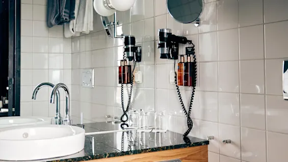 Sink with round basin set in a wooden counter, surrounded by modern bathroom fixtures and white tiled walls; nearby, a toilet with a towel on the lid and a trash bin.