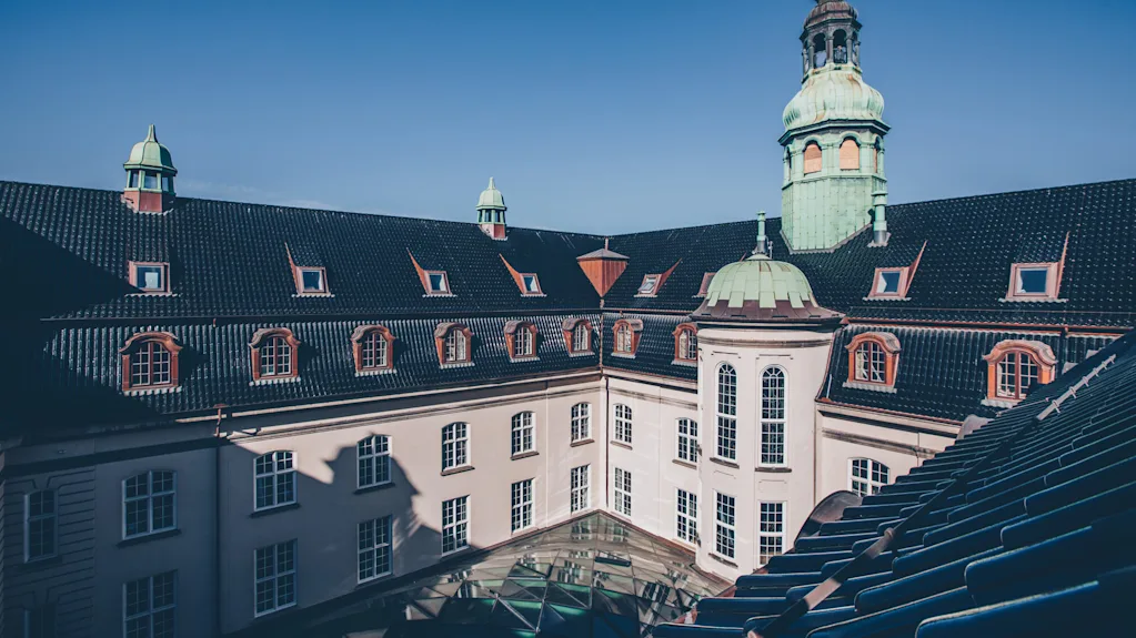 En udsmykket historisk bygning viser en grøn kuppel og tårn mod en klar blå himmel, omgivet af flere vinduer og et mørkt, flisetag.