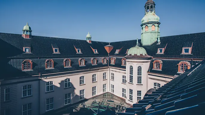 An ornate historical building displays a green dome and tower against a clear blue sky, surrounded by multiple windows and a dark, tiled roof.