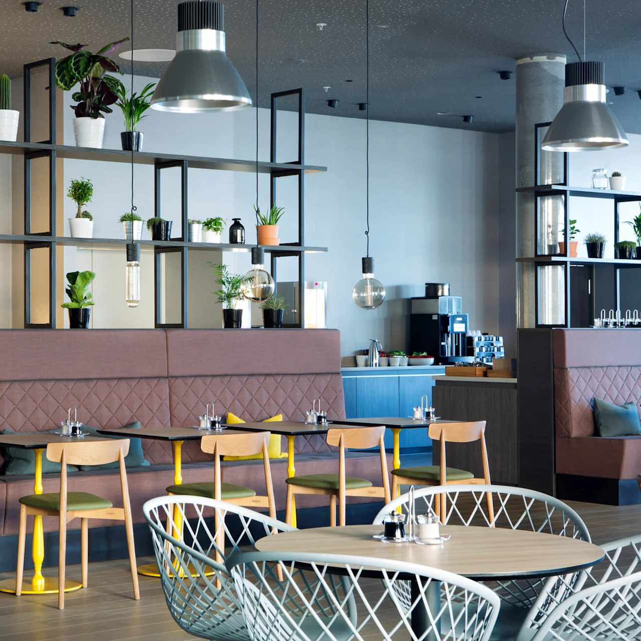Restaurant seating features wooden chairs and tables, set against quilted booths. Hanging plants and light fixtures decorate the space, with a coffee station visible in the background.