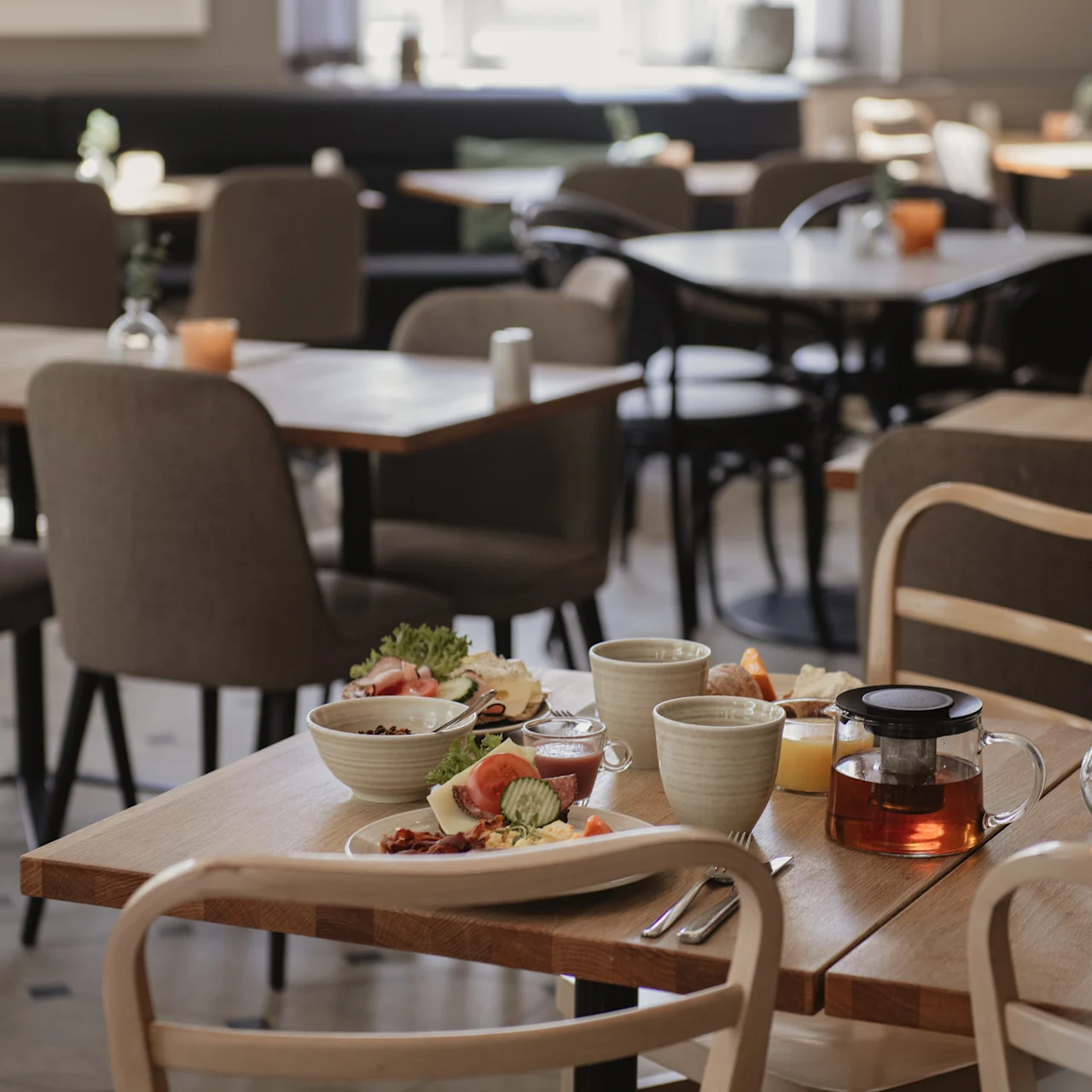 Ett bord med frukostvaror—sallad, skivat bröd, te och koppar—står i ett mysigt, solbelyst café med trästolar och bord runtomkring.