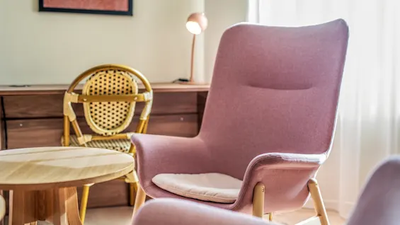 Pink lounge chair sits by a wooden table in a modern, cozy living room. A wicker chair and sideboard sit nearby under soft lighting from a lamp.