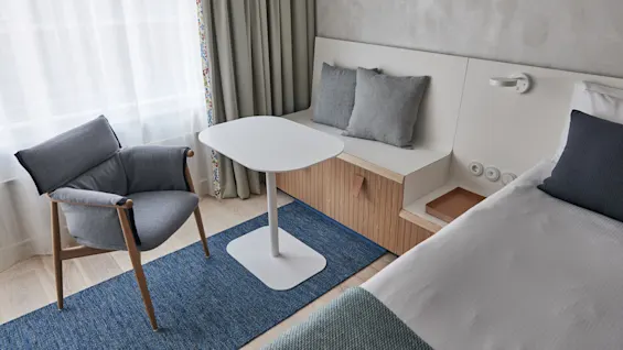 A gray armchair sits beside a white table on a blue rug in a modern, minimalist hotel room with a bed, textured wall, curtains, and a built-in wooden bench.