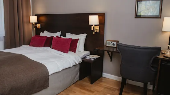 En seng med hvide lagner og røde puder hviler mod et træhovedgærde med lamper, ved siden af et skrivebord med en stol i et tæppebelagt hotelværelse.