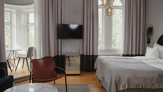 Ett sovrum har en säng med grå sängkläder, riktad mot en väggmonterad TV. I närheten står en röd stol som vetter mot ett bord med marmorskiva. Stora fönster ger naturligt ljus och utomhusutsikt.