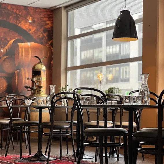 Bord uppsatta med glas i ljuset av svarta hänglampor, omgivna av stolar. En mural med vintunnor, stora fönster och växter i bakgrunden skapar en mysig restaurangatmosfär.