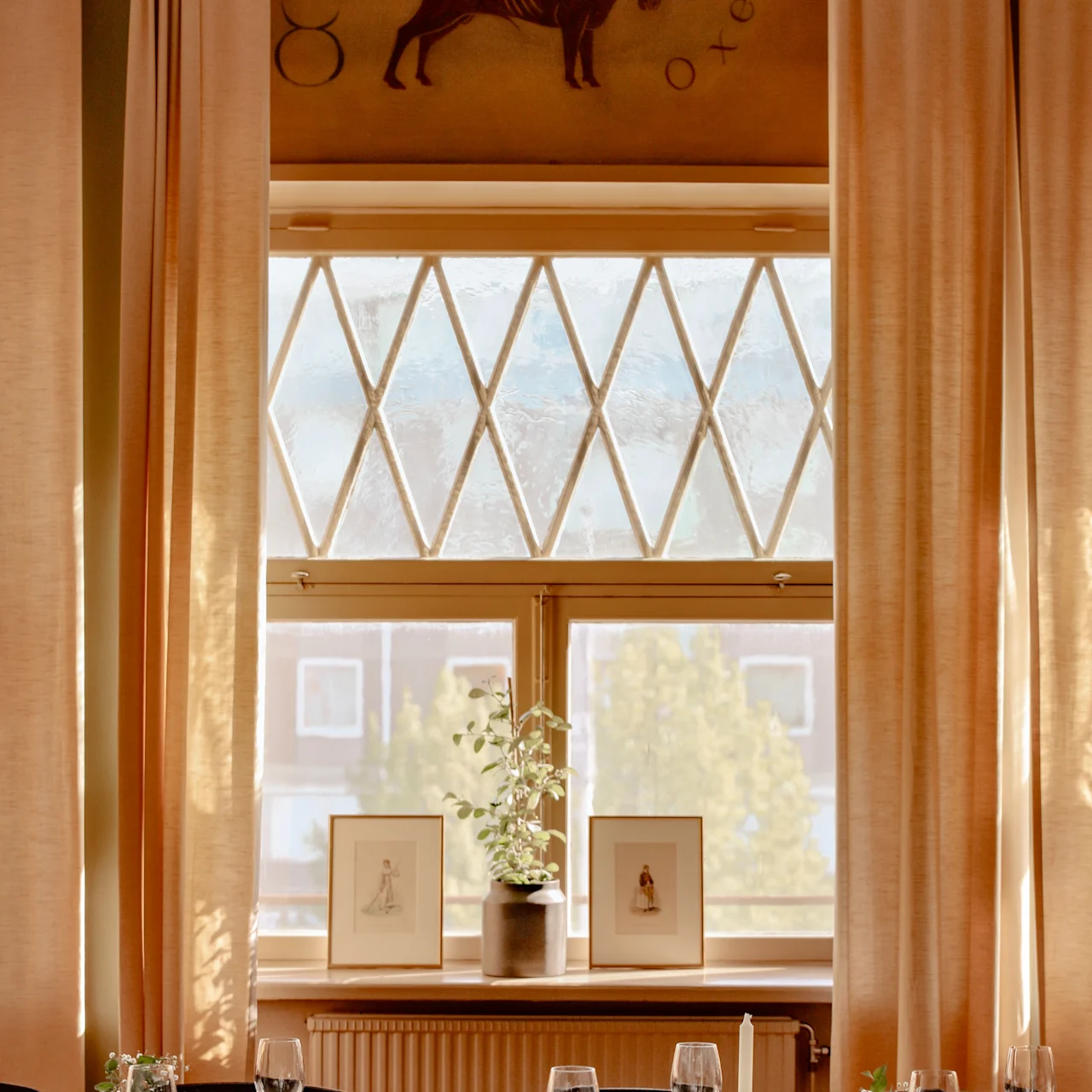 A dining table is set elegantly with glasses and cutlery; behind, a window with beige curtains displays framed art. Above is a mural of a bull with "Oxen" written.
