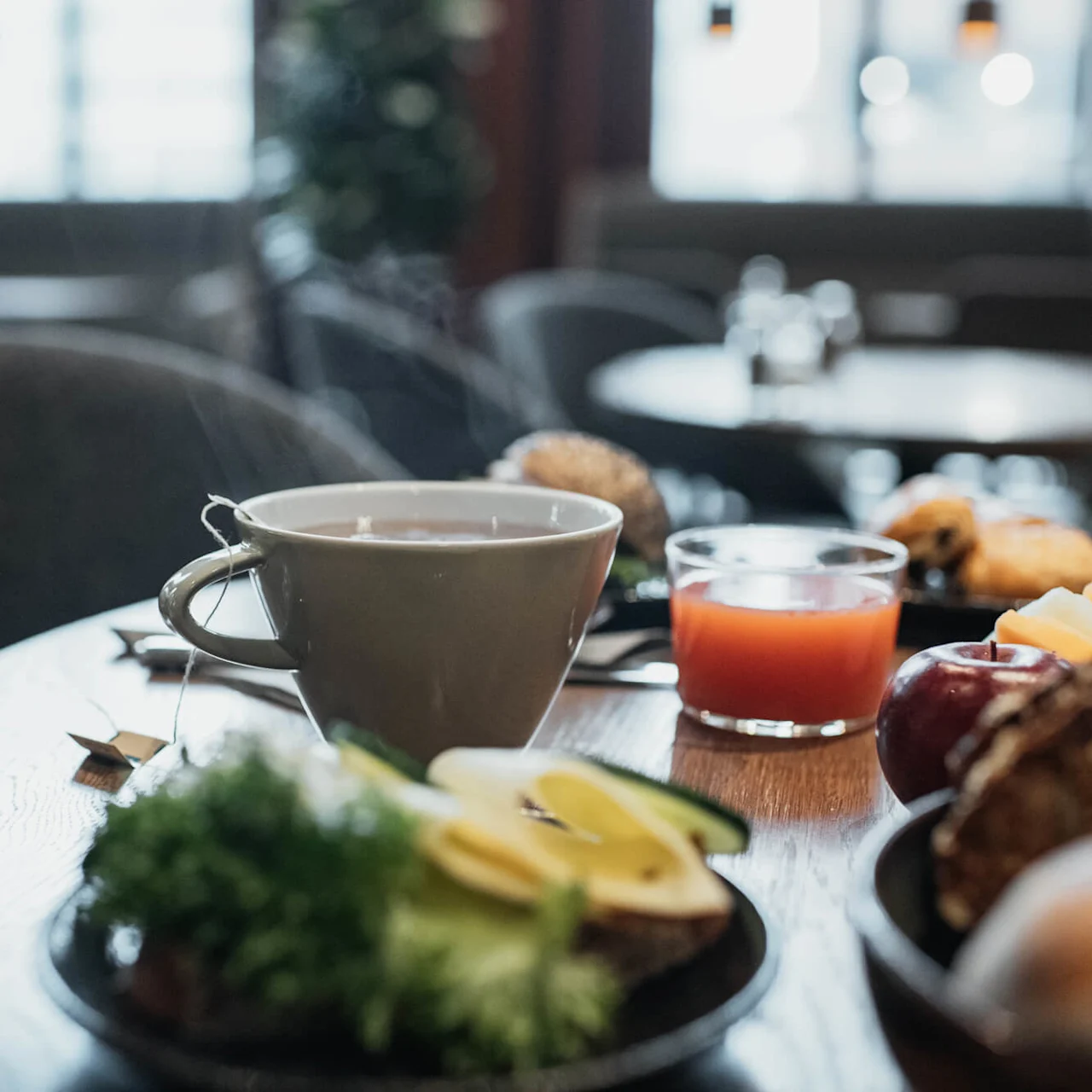 A steaming cup of tea with breakfast items on a wooden table, in a cozy cafe setting.
