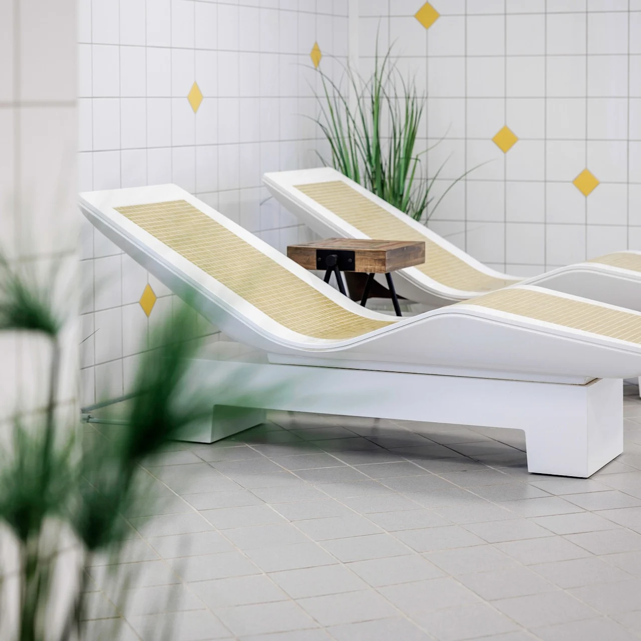 Two white lounge chairs with bamboo inserts rest on a tiled floor beside a small wooden table, surrounded by plants and yellow-diamond patterned white tiles on the walls.